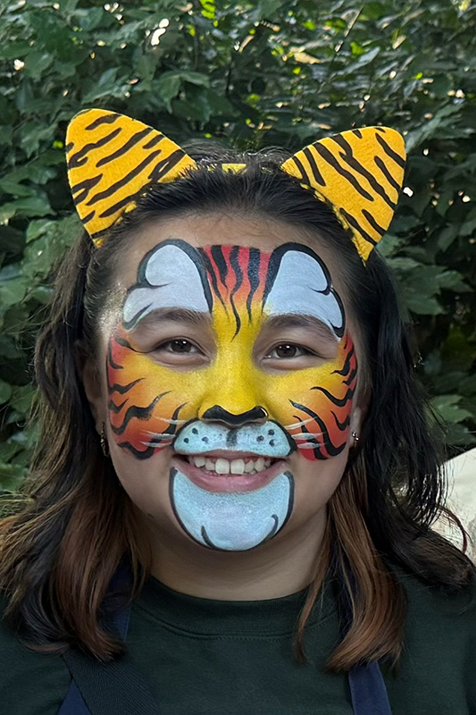 A smiling person with face paint resembling a tiger, featuring yellow, orange, white, and black patterns, and wearing yellow tiger-striped ears headband. Green foliage is visible in the background.