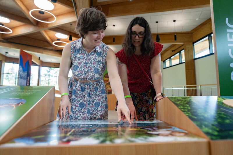 Two women stand indoors at an interactive display, both pointing and smiling as they look at information on the exhibit. The modern room has wooden beams and large windows.