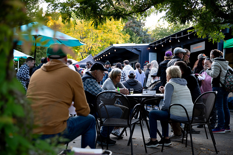 People sit at outdoor tables and chat while others stand in groups near food stalls under trees and umbrellas at a bustling outdoor event on a sunny day.