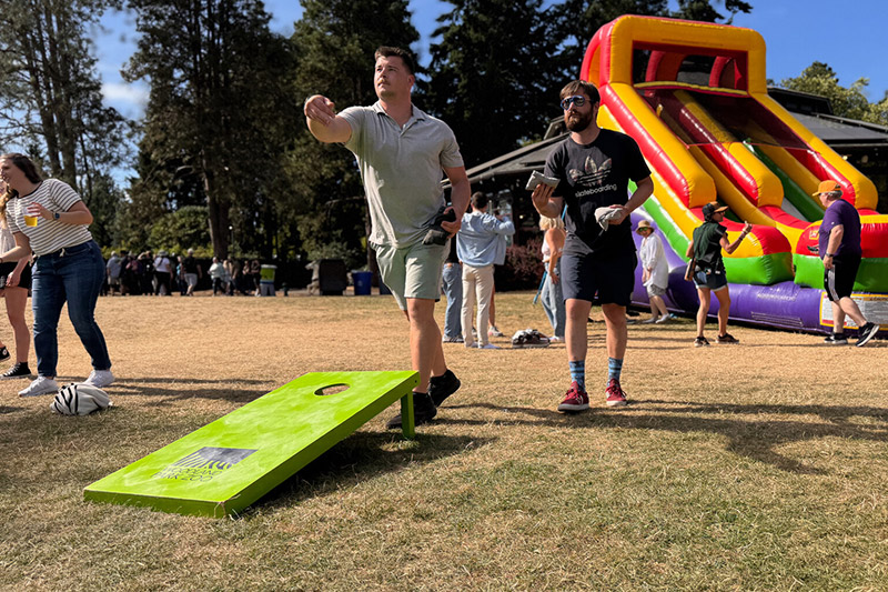 Two people play cornhole on a grassy field at an outdoor event, with others nearby and a colorful inflatable slide in the background.