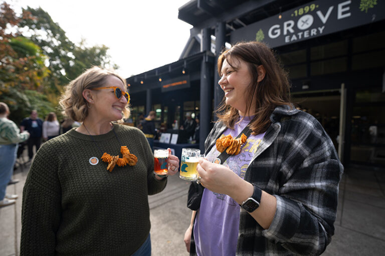 Two women smile and hold drinks outside the 1899 Grove Eatery. Both wear pretzel necklaces. People and trees are visible in the background on a sunny day.