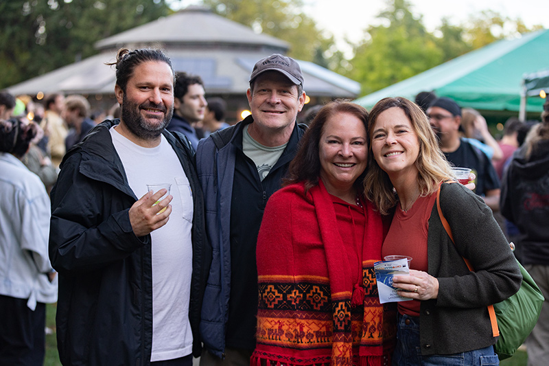 Four adults stand close together smiling at an outdoor event. Two hold drinks. People and tents are visible in the background.